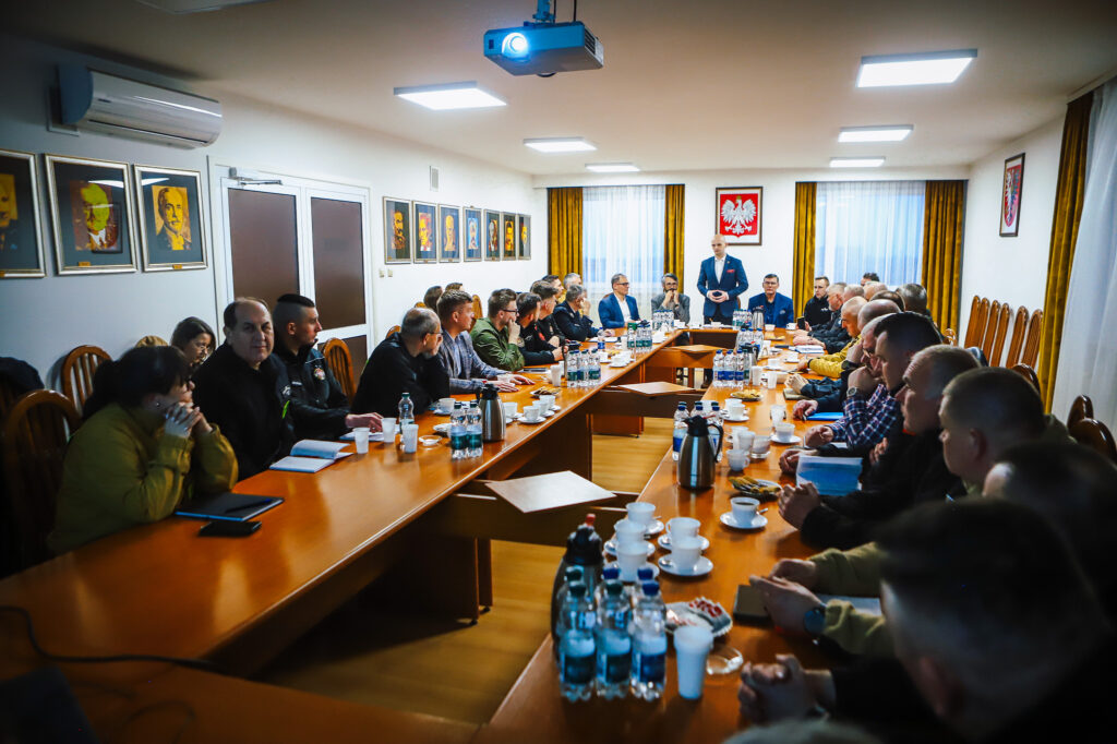 Strażacy ochotnicy spotykają się z przedstawicielami Fundacji ORLEN podczas spotkania w sali konferencyjnej, a stojący na końcu sali mężczyzna wygłasza przemówienie.