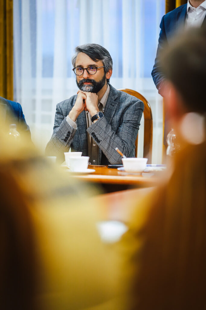 Mężczyzna w kraciastym garniturze siedzi przy stole podczas spotkania strażaków ochotników z przedstawicielami Fundacji ORLEN.