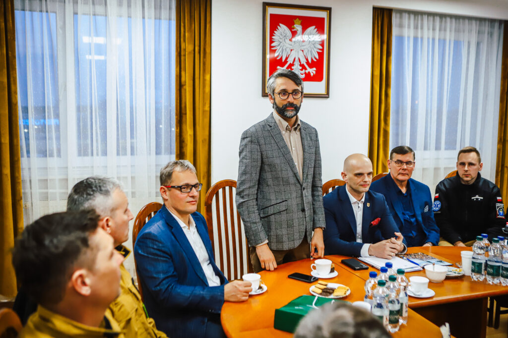 Grupa strażaków ochotników i przedstawicieli Fundacji ORLEN spotyka się przy stole w sali konferencyjnej; na środku stoi mężczyzna w szarej kratkowanej marynarce, a na ścianie widnieje godło Polski.