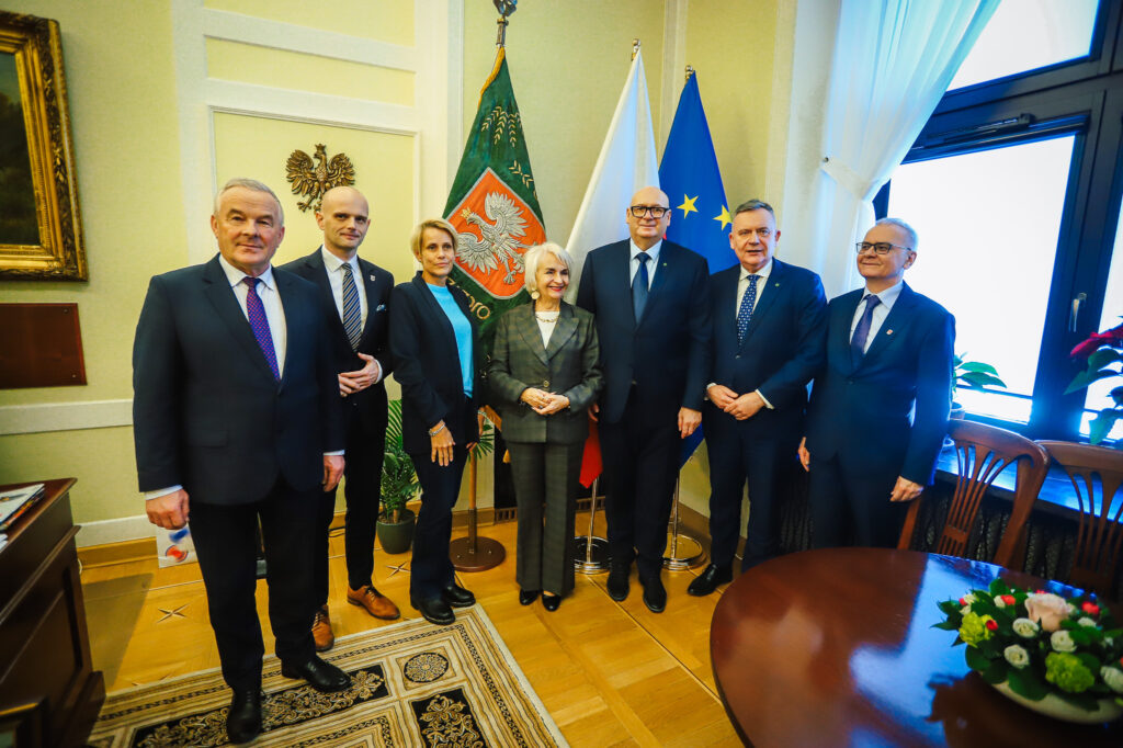 Siedem osób w garniturach stoi w eleganckim pomieszczeniu, na tle flag Polski, Unii Europejskiej i flagi regionalnej.