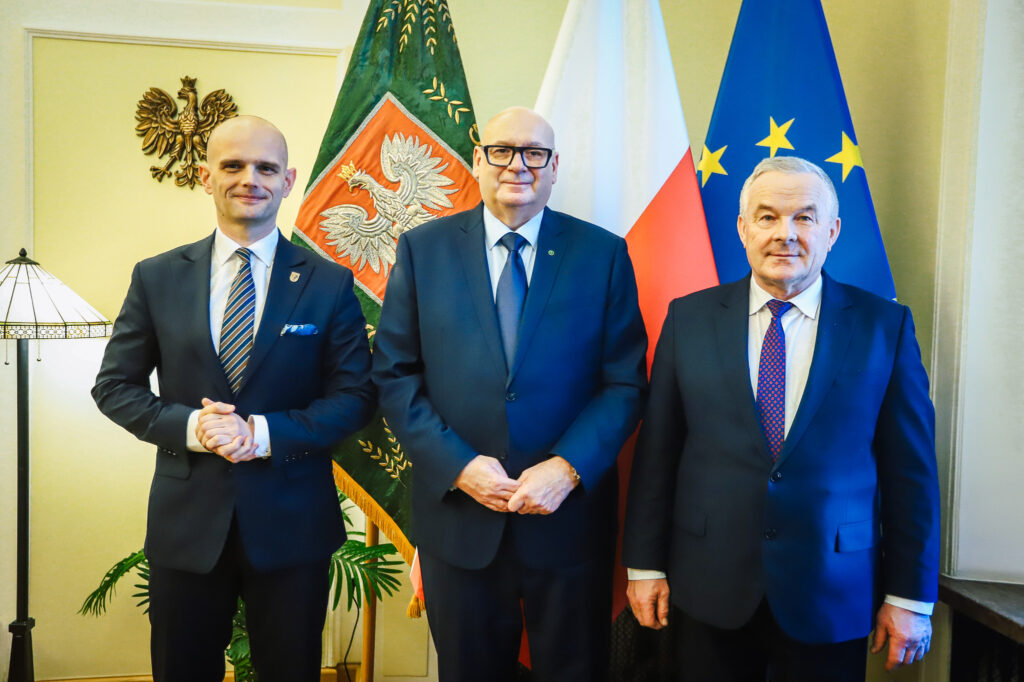 Trzech mężczyzn w garniturach stoi razem w oficjalnym wnętrzu, na tle flag Polski i Unii Europejskiej.