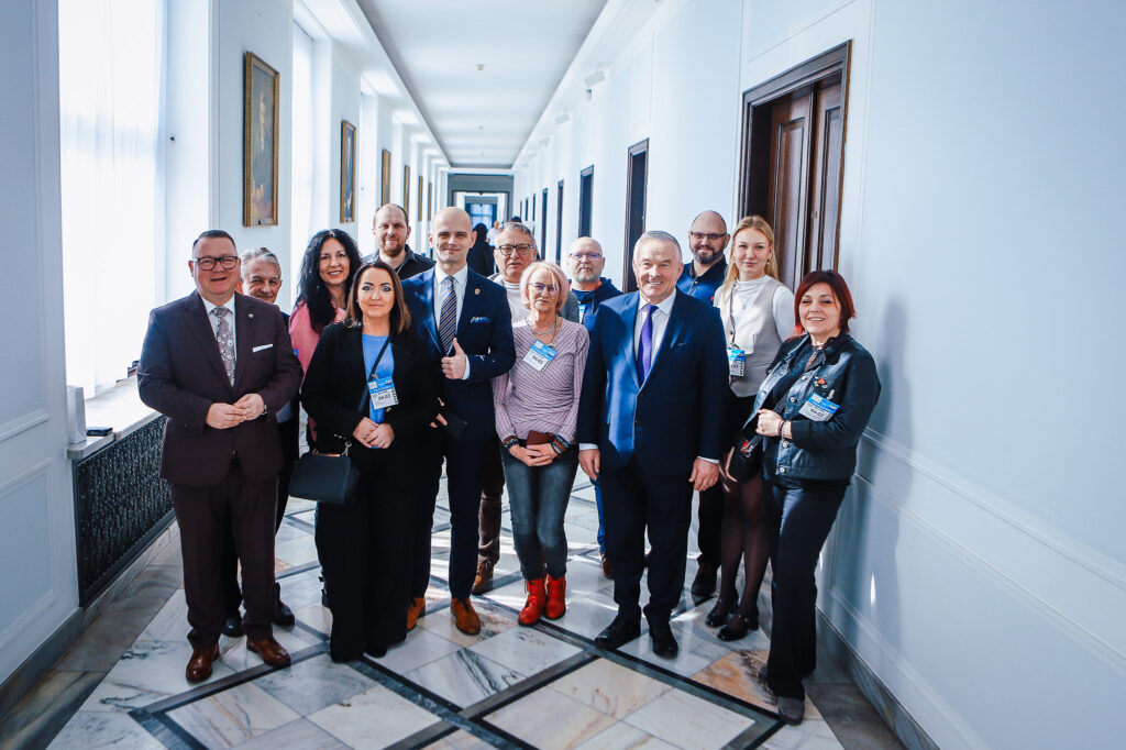 Grupa kilkunastu osób w formalnych strojach stoi razem w jasnym, długim korytarzu z identyfikatorami na szyjach, podczas spotkania dotyczącego planowanej trasy S10 w pobliżu Płocka.