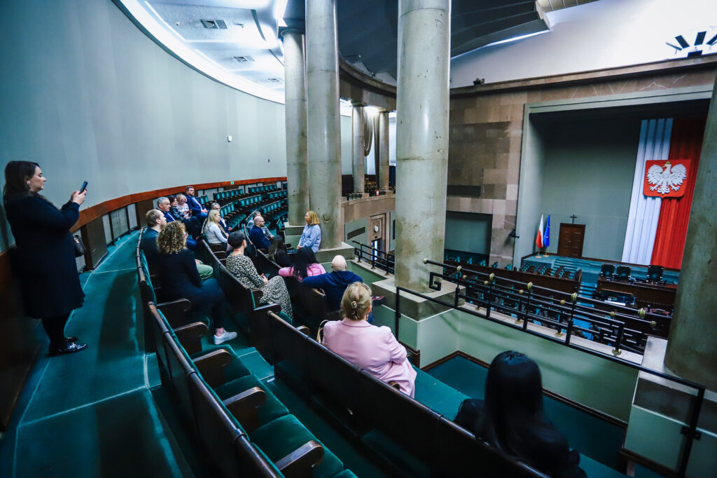 Sala obrad z zielonymi siedzeniami i kilkoma osobami, z wysokimi kolumnami oraz sceną z flagami Polski i Unii Europejskiej i godłem Polski.