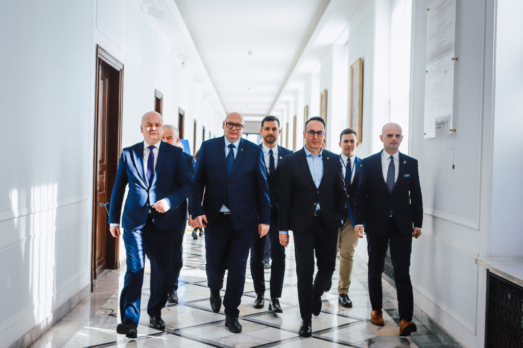 Grupa mężczyzn w garniturach spaceruje korytarzem eleganckiego budynku, odnosząc się do planowanej trasy S10 w pobliżu Płocka.