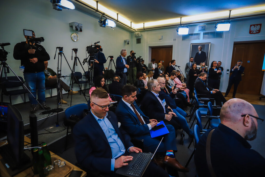 Grupa uczestników konferencji prasowej w sali z kamerami i dziennikarzami, omawiająca pewność przebiegu trasy S10 w pobliżu Płocka.