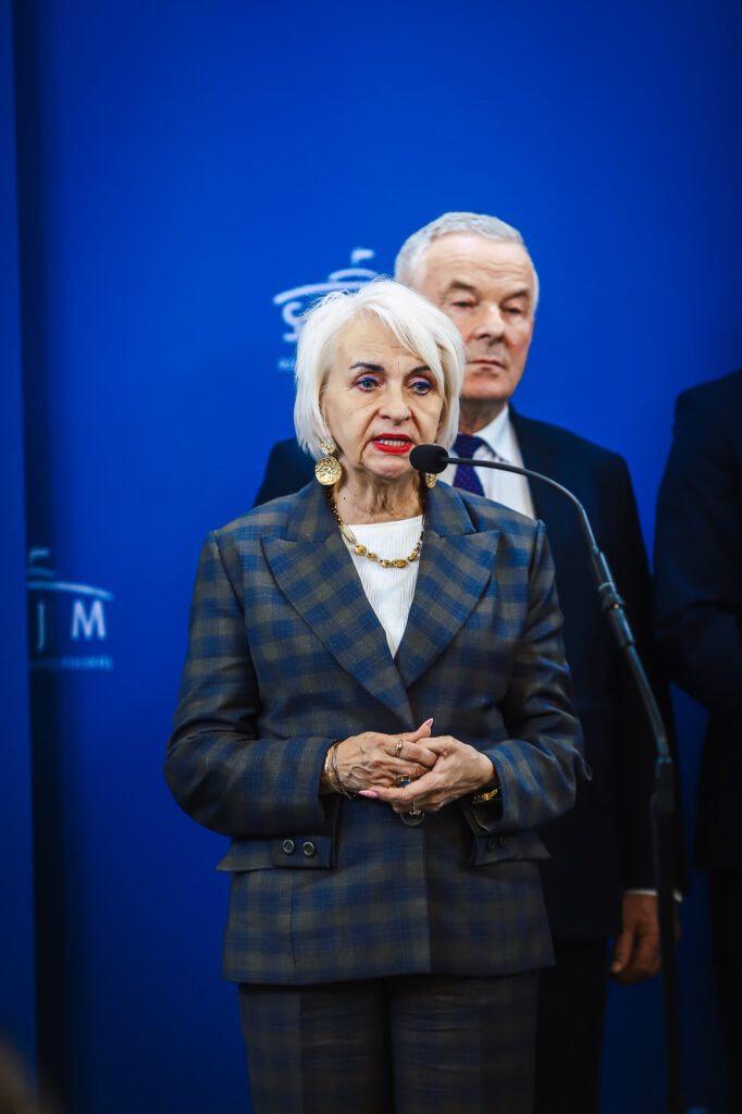 Kobieta w garniturze przemawia do mikrofonu podczas konferencji, na tle niebieskiego tła z logo, w kontekście planowanej trasy S10 przebiegającej w pobliżu Płocka.