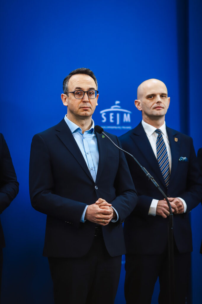 Dwóch mężczyzn w garniturach stoi przy mikrofonie podczas konferencji na niebieskim tle z logo Sejmu.