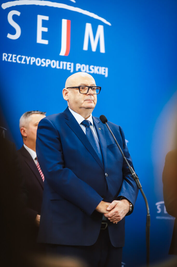 Mężczyzna w granatowym garniturze stoi przy mikrofonie na niebieskim tle z napisem Sejm Rzeczypospolitej Polskiej, podczas konferencji prasowej o planowanej trasie S10 w pobliżu Płocka.