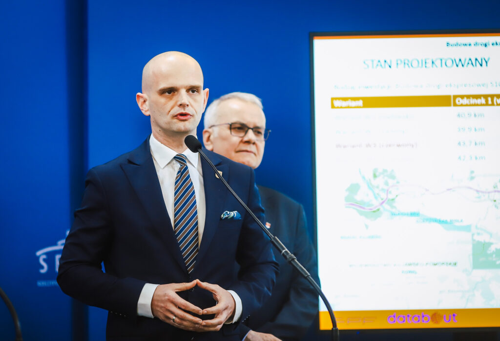 Dwóch mężczyzn w garniturach przemawia przy mikrofonie na konferencji prasowej, obok ekranu z prezentacją planowanej trasy S10 w pobliżu Płocka.
