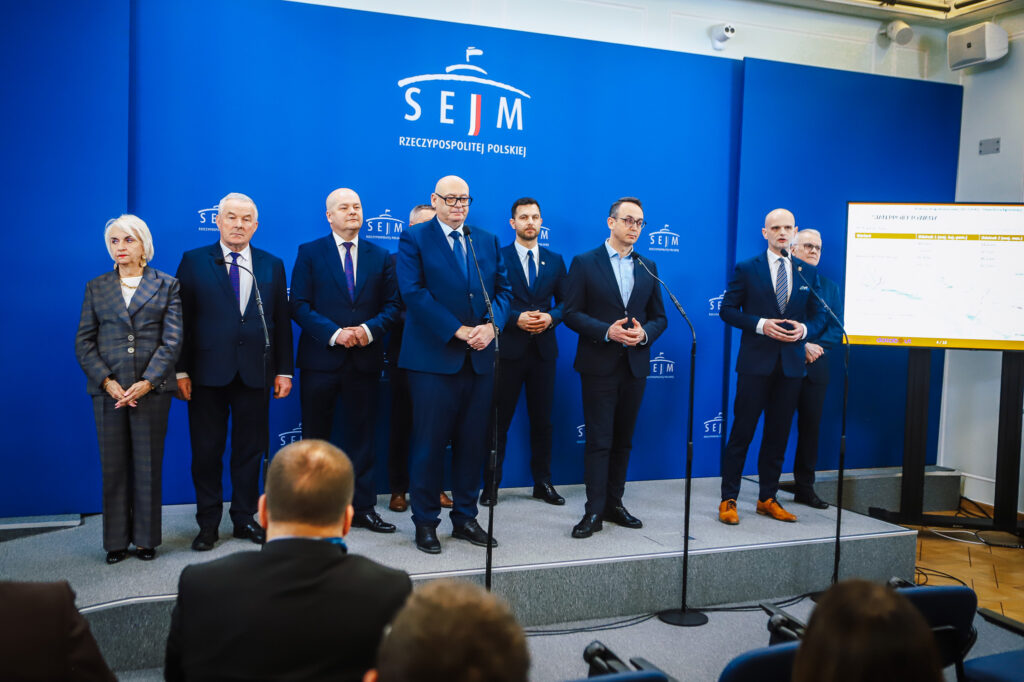 Grupa polityków stojących na scenie podczas konferencji w Sejmie, na tle niebieskiego loga Sejmu, omawia planowaną trasę S10 w pobliżu Płocka.