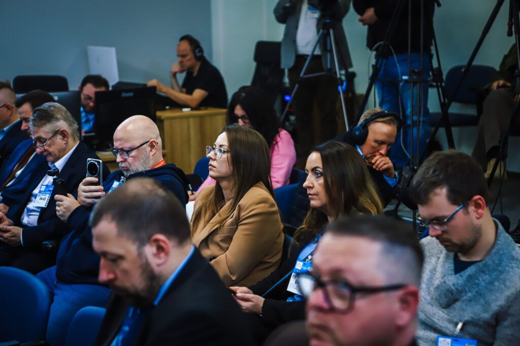 Uczestnicy konferencji prasowej siedzą w sali, mają identyfikatory na szyjach, a w tle widać ekipę filmową i sprzęt reporterski.