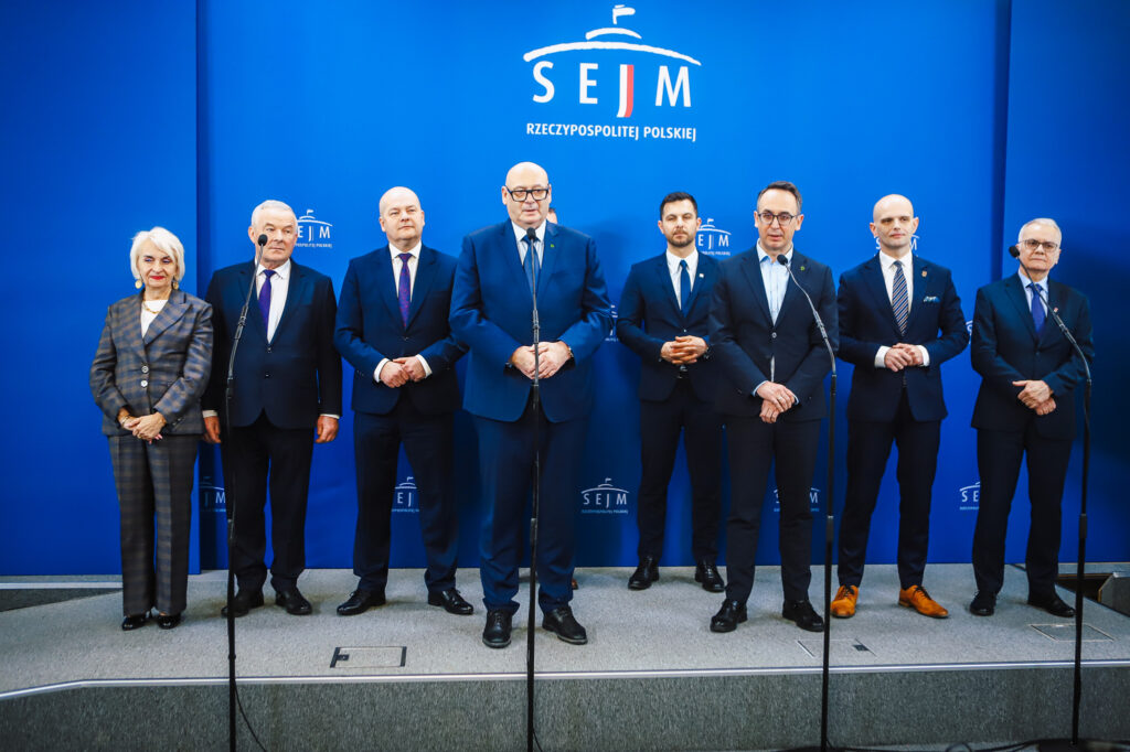 Grupa polityków w garniturach stoi na scenie przed niebieską ścianką z logo Sejmu podczas konferencji prasowej dotyczącej przebiegu trasy S10 w pobliżu Płocka.