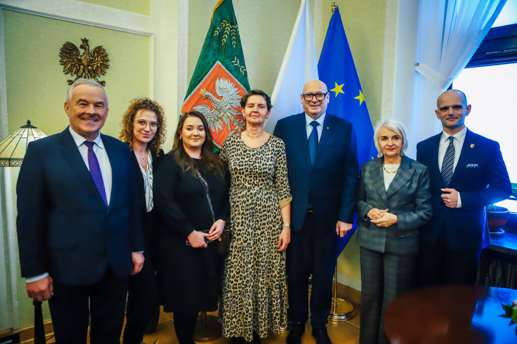 Siedmioosobowa grupa osób w formalnych ubraniach stoi w biurze, za nimi widoczne flagi UE i flagi regionalne, co nawiązuje do planowanej trasy S10 przebiegającej w pobliżu Płocka.