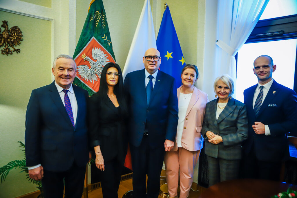 Sześć osób w eleganckich garniturach i marynarkach stoi razem na tle flag państwowych i unijnych w oficjalnym pomieszczeniu.