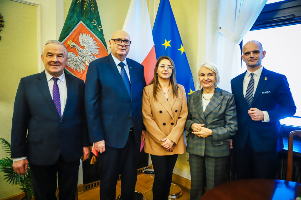 Pięć osób w eleganckich ubraniach stoi razem na tle flag Polski i Unii Europejskiej, podczas wydarzenia dotyczącego planowanej trasy S10 w pobliżu Płocka.