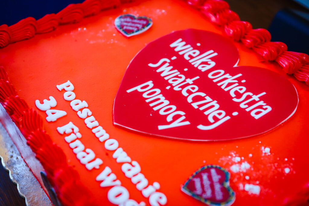 Duży tort w czerwono-pomarańczowych barwach z czerwonym sercem z napisem „Wielka Orkiestra Świątecznej Pomocy” oraz białym napisem „Podsumowanie WOŚP 34 Finał”.