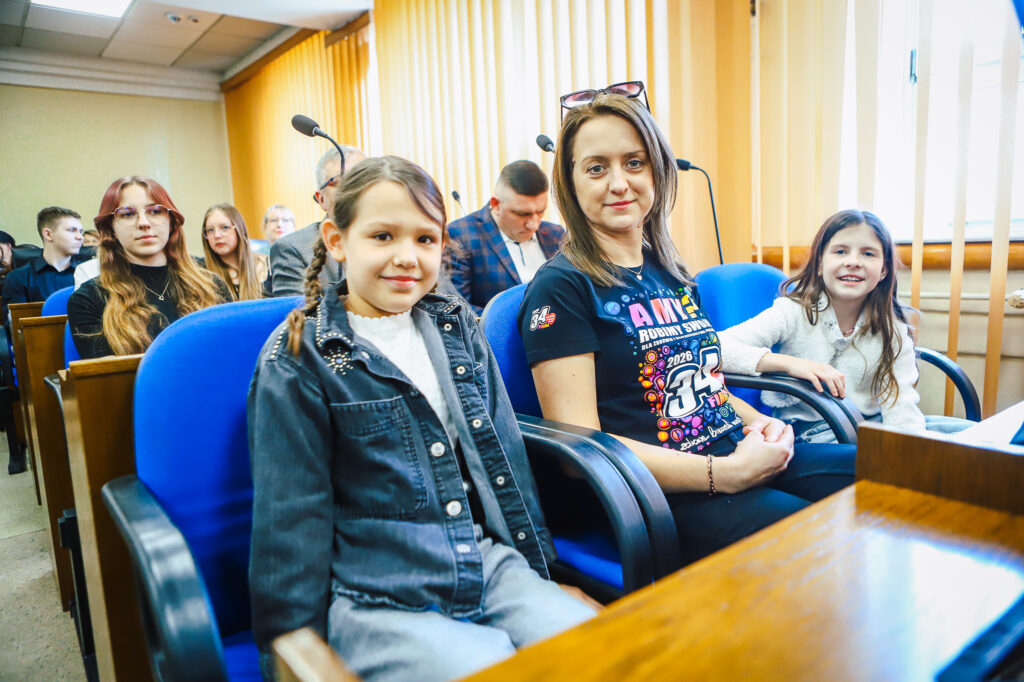 Uczestnicy uroczystego podsumowania Finału WOŚP w powiecie płockim siedzą w sali konferencyjnej na niebieskich fotelach; na pierwszym planie trzy osoby – dwie dziewczynki i dorosła kobieta – wśród innych uczestników i mikrofonów w tle.