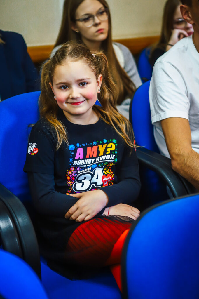 Uśmiechnięta dziewczynka siedzi na niebieskim krześle podczas uroczystego podsumowania tegorocznego Finału WOŚP w powiecie płockim.