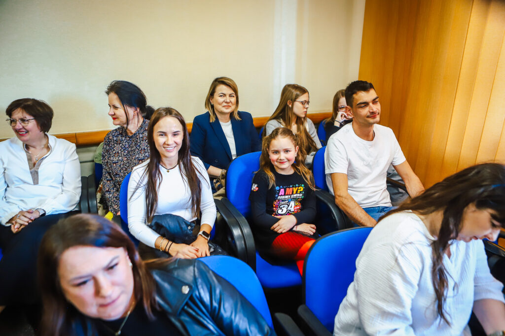 Uczestnicy uroczystego podsumowania tegorocznego Finału WOŚP w powiecie płockim siedzą na niebieskich krzesłach w sali konferencyjnej.