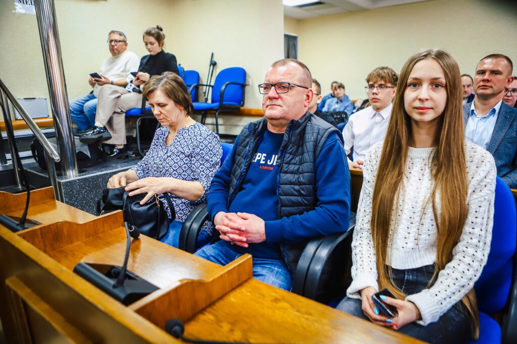 Uczestnicy uroczystego podsumowania tegorocznego Finału WOŚP w powiecie płockim siedzą w sali; na pierwszym planie kobieta otwierająca torebkę, mężczyzna w niebieskiej bluzie i młoda dziewczyna z długimi włosami w jasnym swetrze.