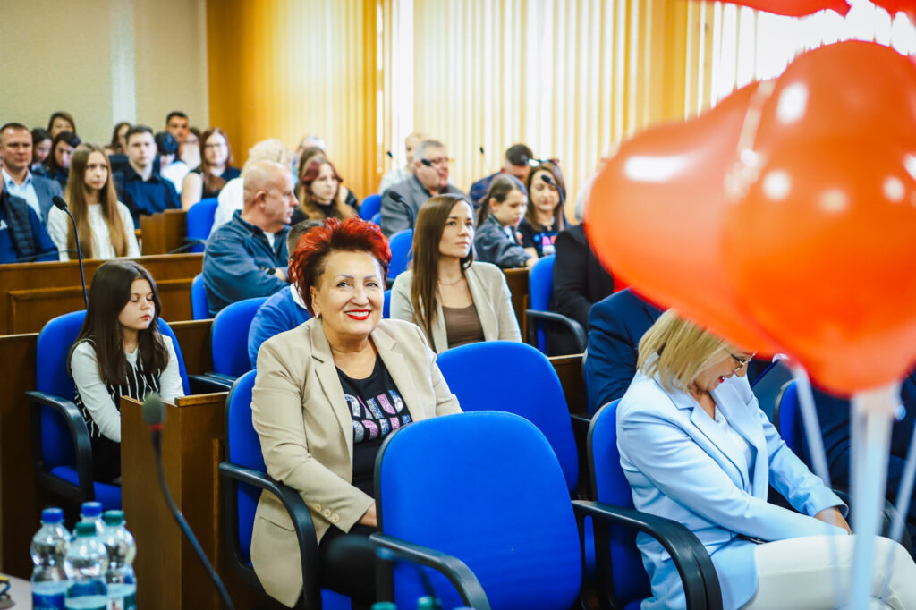 Uczestnicy uroczystego podsumowania Finału WOŚP w powiecie płockim siedzą w sali z niebieskimi fotelami; na pierwszym planie uśmiechnięta kobieta o czerwonych włosach, a po prawej widoczny czerwony balon w kształcie serca.