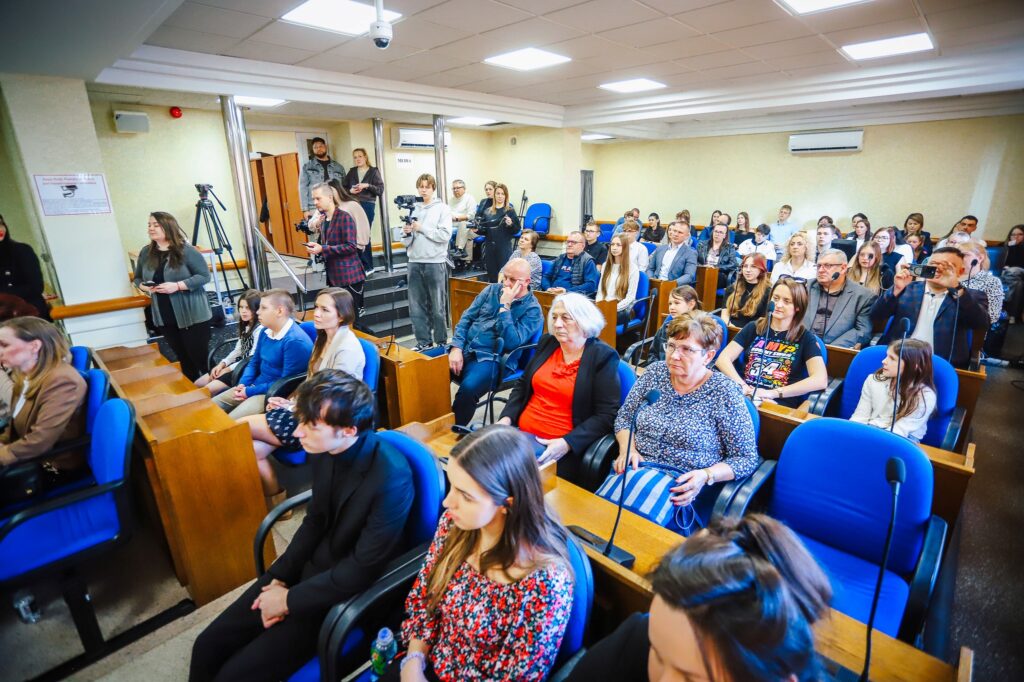 Uczestnicy uroczystości podsumowania tegorocznego Finału WOŚP w powiecie płockim siedzą w sali konferencyjnej, a obok nich stoją dziennikarze z kamerami.