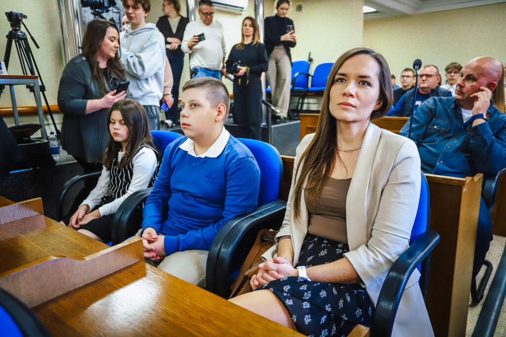 Grupa uczestników siedzi w sali podczas uroczystego podsumowania tegorocznego Finału WOŚP w powiecie płockim; na pierwszym planie kobieta w beżowym żakiecie, chłopiec w niebieskim swetrze i dziewczynka w czarno-białej sukience.