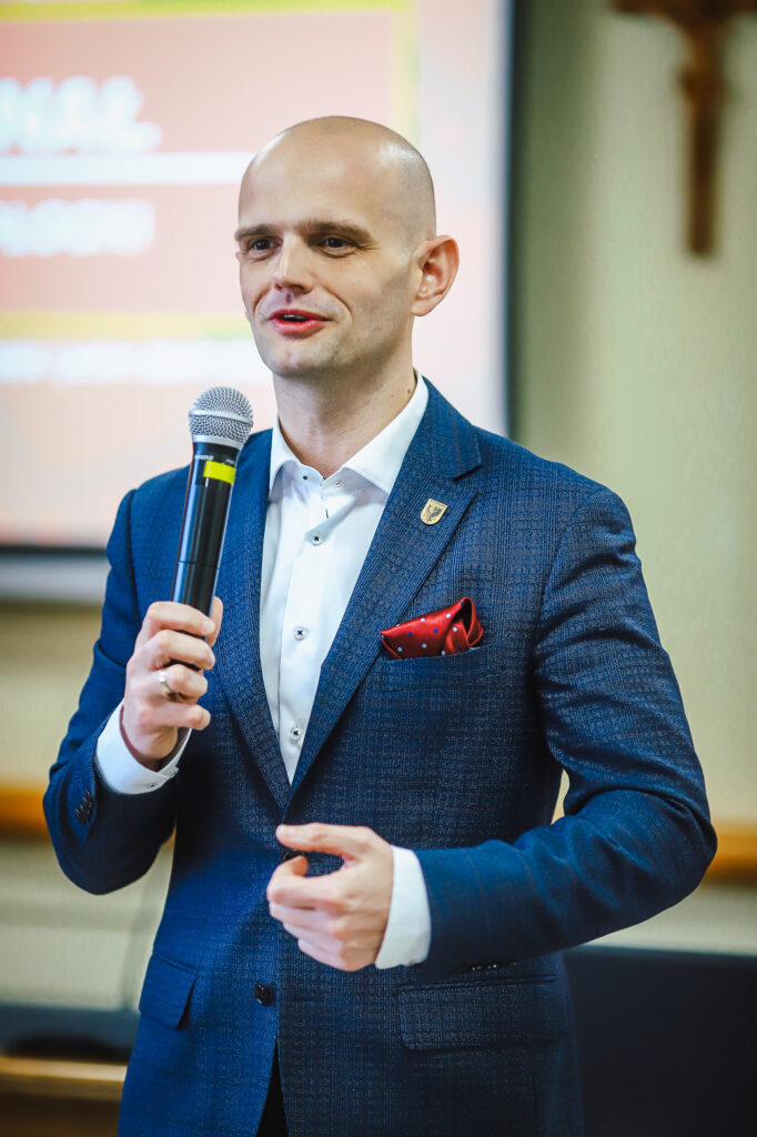 Mężczyzna w granatowym garniturze z czerwonym poszetką w kieszeni przemawia do mikrofonu podczas uroczystego podsumowania Finału WOŚP w powiecie płockim.