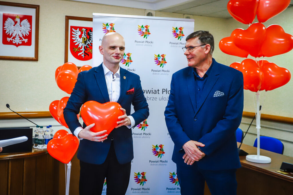 Dwóch mężczyzn w garniturach stoi na tle banneru z logotypem Powiatu Płockiego, trzymając czerwone balony w kształcie serc podczas uroczystości podsumowania Finału WOŚP w powiecie płockim.