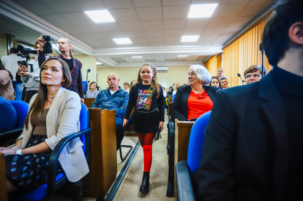 Dziewczynka w czarnym stroju z czerwonymi rajstopami spaceruje środkiem sali podczas uroczystego podsumowania tegorocznego Finału WOŚP w powiecie płockim, wśród zgromadzonych widzów i kamer.