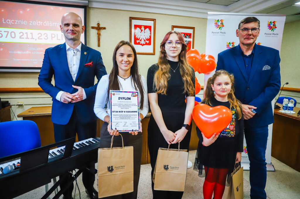 Pięć osób stoi na scenie podczas uroczystego podsumowania Finału WOŚP w powiecie płockim, trzymając Dyplom i torby z logo powiatu oraz sercowy balon, a w tle widać prezentację z zebranymi pieniędzmi.