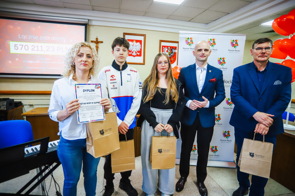 Grupa osób stoi na sali podczas uroczystego podsumowania Finału WOŚP w powiecie płockim, trzymając dyplom i papierowe torby z logo Powiatu Płockiego, a w tle widać ekran z kwotą zebranych pieniędzy.