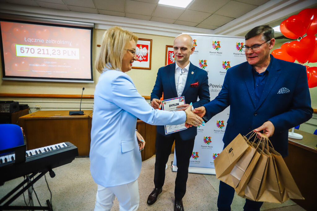 Uroczysta scena podsumowania Finału WOŚP w powiecie płockim: kobieta w jasnoniebieskiej marynarce wręcza dyplom mężczyźnie w granatowym garniturze, obok stoi trzeci mężczyzna z torbami na prezenty, a w tle widać ekran z zebranymi pieniędzmi i banner Powiat Płocki oraz balony.