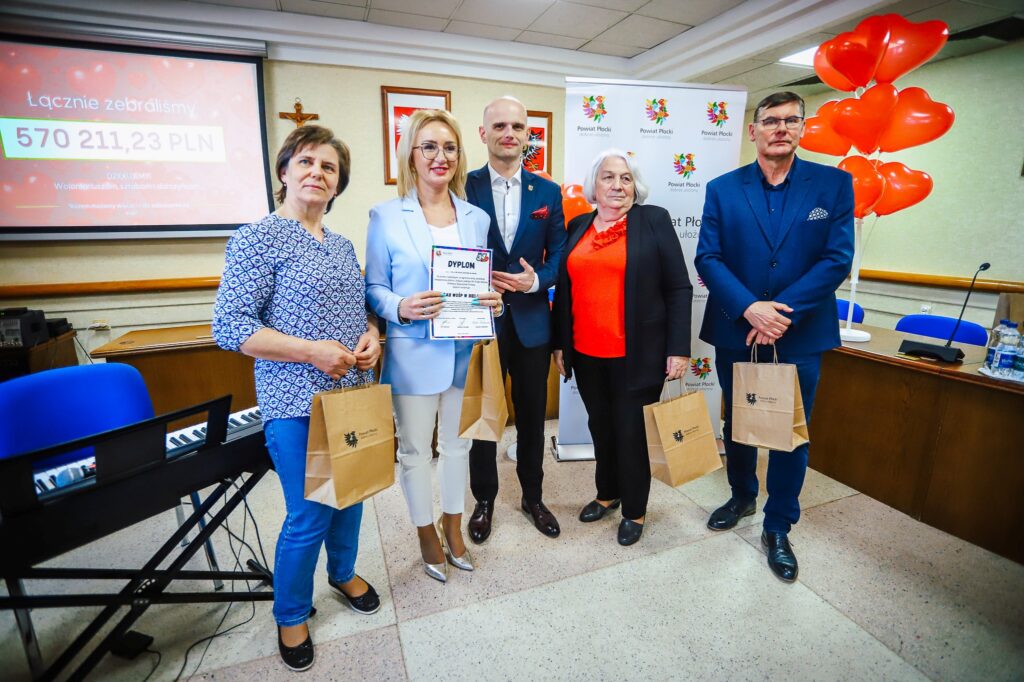 Pięć osób stoi na sali podczas uroczystości podsumowania Finału WOŚP w powiecie płockim, trzymają dyplom i torby z upominkami, a w tle są banery i balony w kształcie serc.