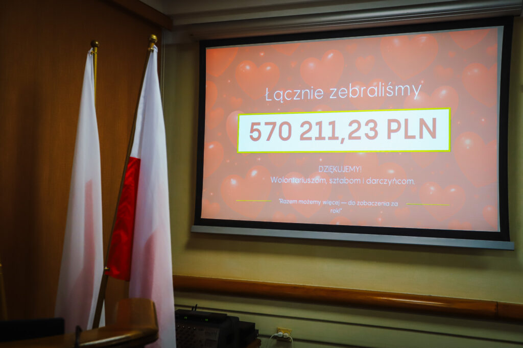 Sala konferencyjna, na ekranie wyświetlono podsumowanie Finału WOŚP w powiecie płockim z kwotą 570 211,23 PLN, a obok stoją flagi.