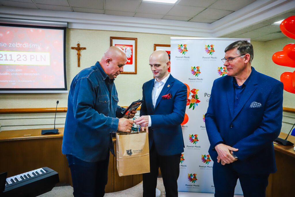 Uroczyste podsumowanie tegorocznego Finału WOŚP w powiecie płockim: mężczyzna w jeansowej koszuli wręcza torbę z logo Powiatu Płockiego innemu uczestnikowi, podczas gdy trzeci obserwuje, na tle banerów i ekranu.