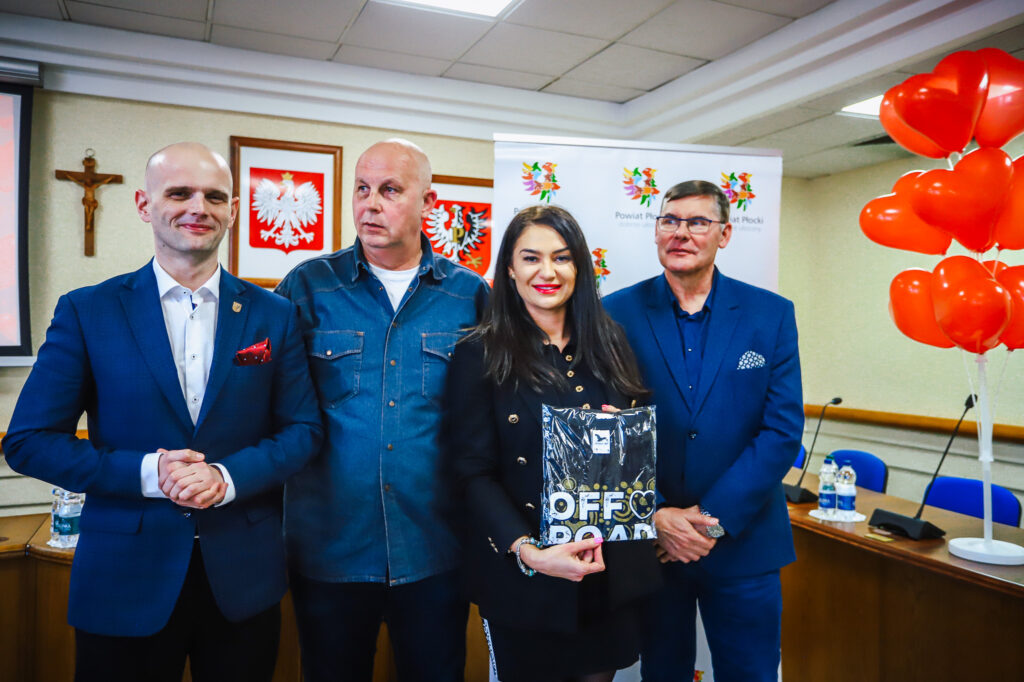 Cztery osoby stoją w sali podczas uroczystego podsumowania Finału WOŚP w powiecie płockim, a w tle widać balony w kształcie serc, polskie godła i baner Powiatu Płockiego.