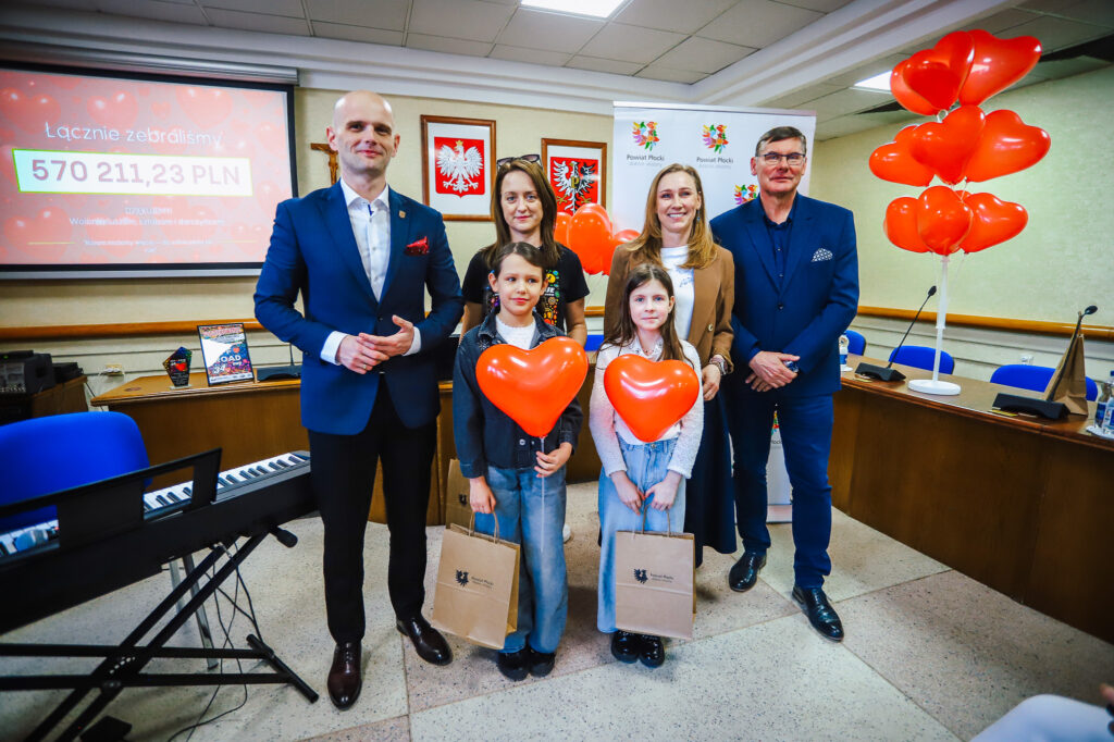 Grupa osób w sali, wśród nich dwie dziewczynki z czerwonymi balonami w kształcie serc, podczas uroczystego podsumowania Finału WOŚP w powiecie płockim; w tle wyświetlana suma 570 211,23 PLN i loga Powiatu Płockiego.