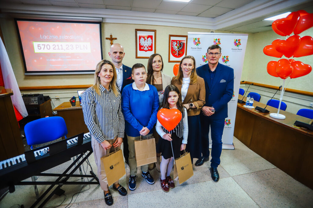 Grupa osób podczas uroczystego podsumowania Finału WOŚP w powiecie płockim, stojąca przed ekranem z informacją o zebranej kwocie 570 211,23 PLN i czerwonymi balonami w kształcie serc.