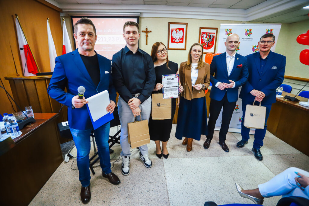 Sześć osób stoi w sali konferencyjnej z flagami i godłami, trzyma dyplom i torby z upominkami podczas uroczystości podsumowania Finału WOŚP w powiecie płockim.