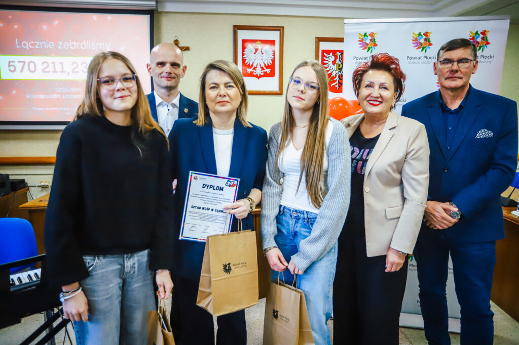 Grupa sześciu osób na sali podczas uroczystości podsumowania Finału WOŚP w powiecie płockim, trzymająca dyplom i torby z logo Powiatu Płockiego na tle prezentacji i banerów.