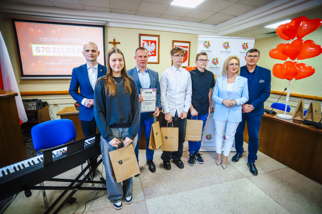 Grupa osób stoi w sali podczas uroczystego podsumowania tegorocznego Finału WOŚP w powiecie płockim, trzymając dyplom i torby z upominkami, a w tle widać prezentację z kwotą zebranych pieniędzy i czerwone balony w kształcie serc.