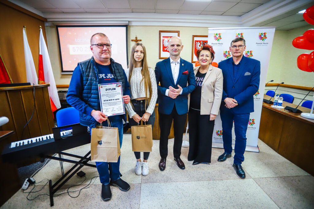 Pięć osób stoi na sali podczas uroczystego podsumowania Finału WOŚP w powiecie płockim, trzymając dyplom i torby z prezentami, na tle flag, ekranu i banerów Powiatu Płockiego.