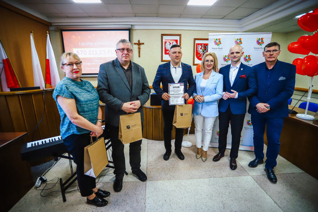Sześcioosobowa grupa stoi na sali konferencyjnej Powiatu Płockiego podczas uroczystego podsumowania Finału WOŚP, trzymając dyplom i torby z upominkami, a w tle widać flagi, krzyż i banery.