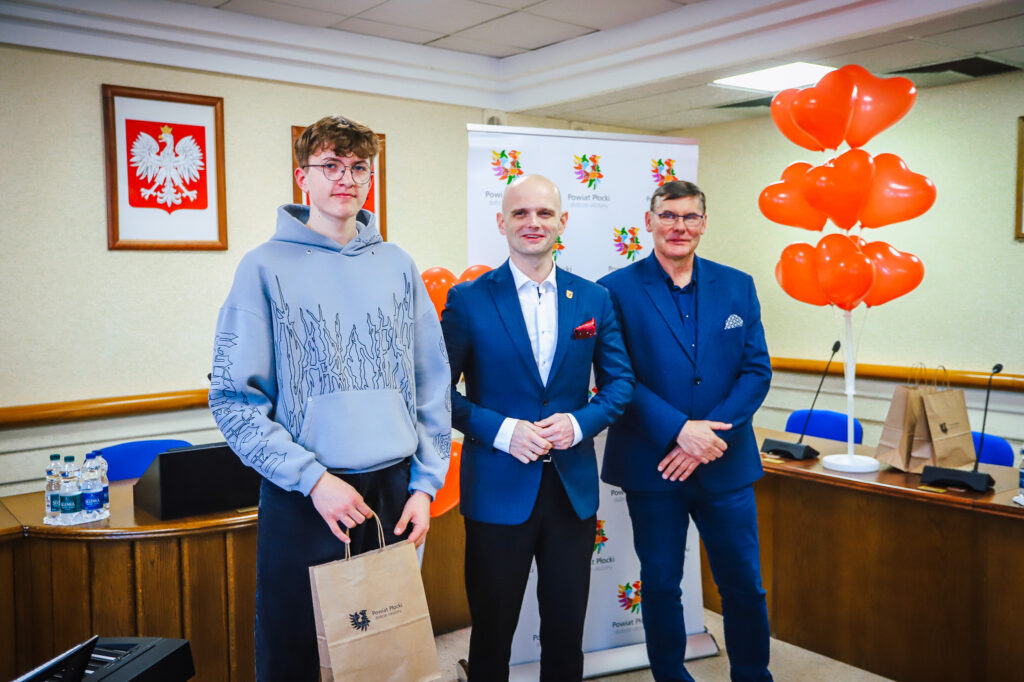 Trzech mężczyzn stoi w sali podczas uroczystego podsumowania Finału WOŚP w powiecie płockim: młody mężczyzna po lewej w bluzie trzyma torbę z logo powiatu, a po środku i po prawej stoją w garniturach, w tle baner Powiat Płocki, balony w kształcie serc i godło Polski na ścianie.