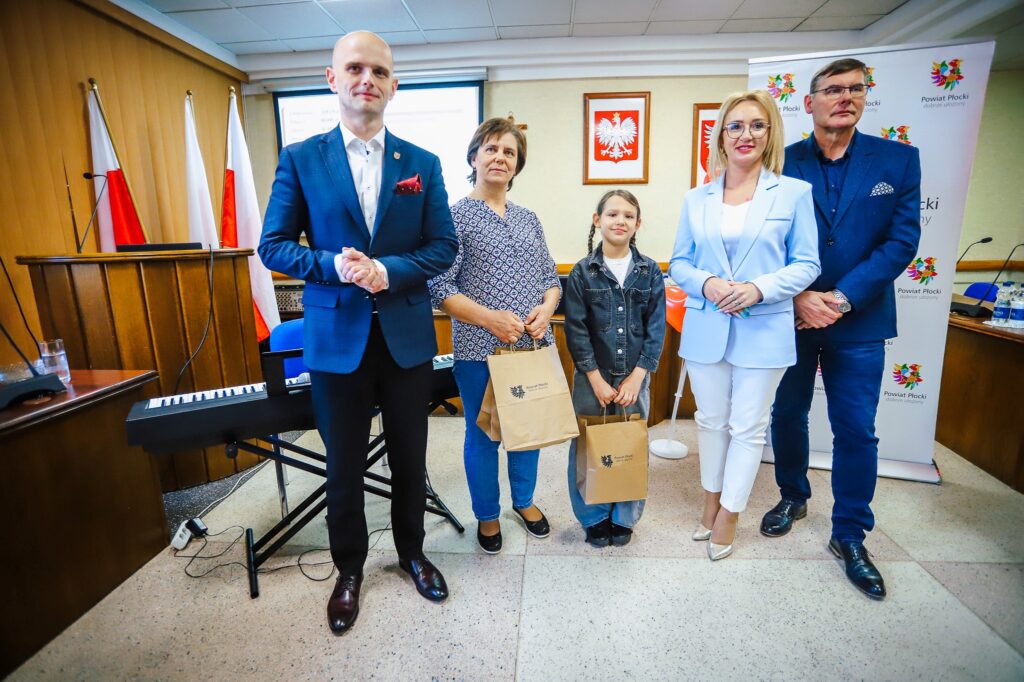 Grupa pięciu osób stoi w sali konferencyjnej podczas uroczystego podsumowania Finału WOŚP w powiecie płockim, trzymając torby z upominkami, obok stoi pianino, a w tle widoczne są flagi państwowe i baner z logotypami Powiatu Płockiego.