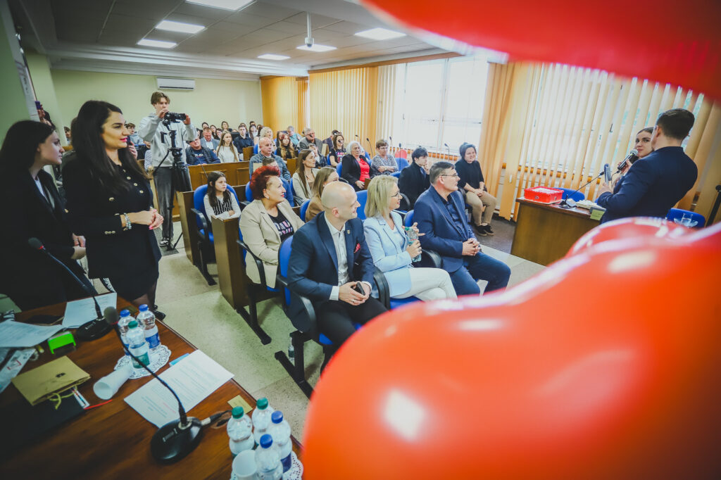 Prowadzący z mikrofonem przemawia na mównicy podczas uroczystego podsumowania tegorocznego Finału WOŚP w powiecie płockim, przed licznie zgromadzoną publicznością w sali.