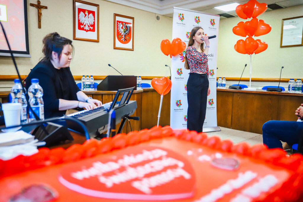 Wokalistka śpiewa z mikrofonem na scenie podczas uroczystego podsumowania Finału WOŚP w powiecie płockim, obok niej gra na keyboardzie muzyk, a w tle widać balony w kształcie serc i banery z herbami powiatu.