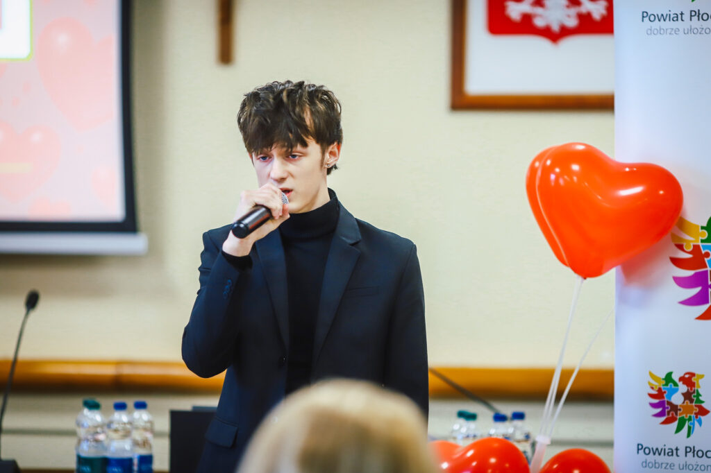 Mężczyzna w czarnym garniturze i czarnym golfie przemawia do mikrofonu podczas uroczystego podsumowania Finału WOŚP w powiecie płockim; na tle widać balony w kształcie serc i baner z logo powiatu.