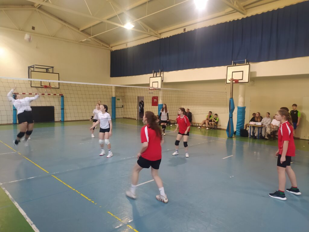 Drużyna dziewcząt w czerwonych i białych koszulkach rozgrywa mecz siatkówki podczas finałów w hali, przy siatce, z widzami siedzącymi na ławkach w tle.